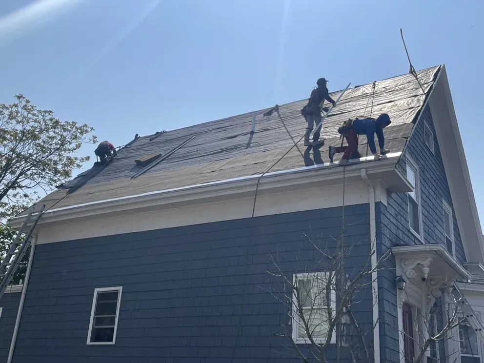 Roof workers repairing shingles on a dark blue house under a clear sky