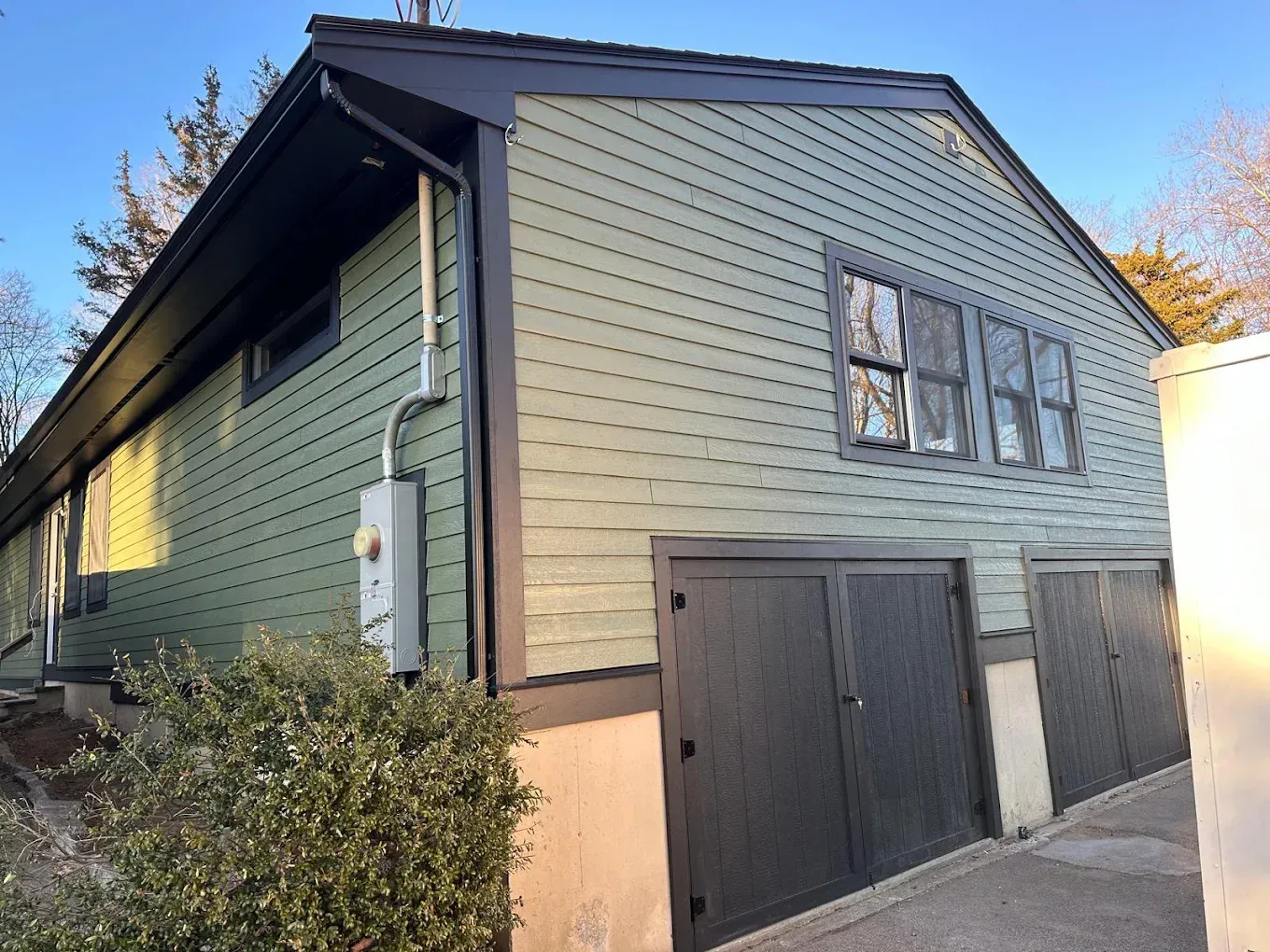 Gray house exterior with dark garage doors, side windows, and bushes along a narrow driveway