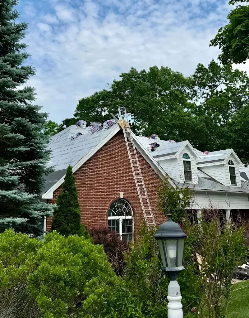 Brick house with white roof trim, arched window, and ladder against lush trees under a cloudy sky