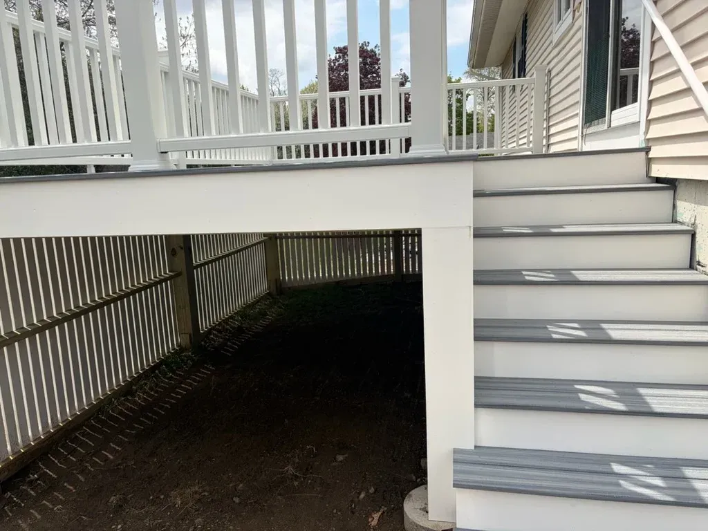 Covered deck area beneath white porch with gray stairs and railing, casting striped shadows on the ground