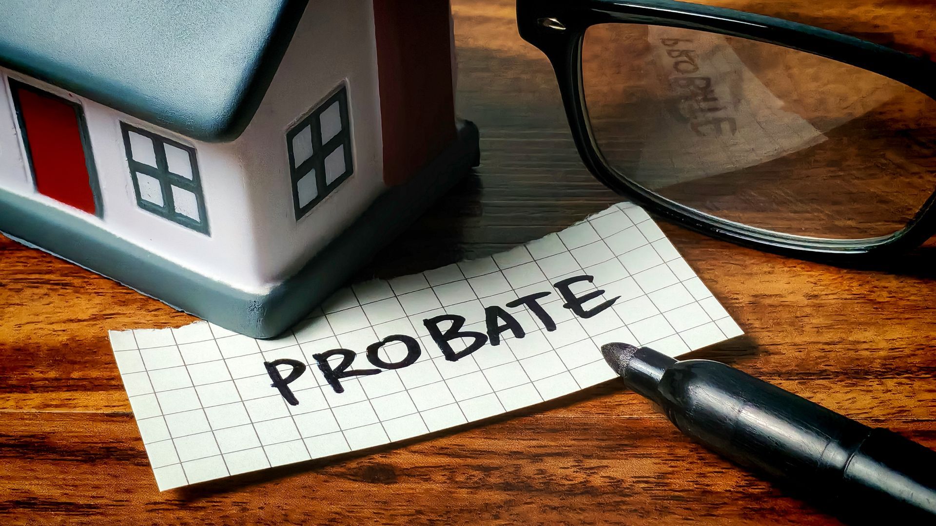 Note labeled probate next to a model house, pen, and glasses on a wooden desk Note labeled probate next to a model house, pen, and glasses on a wooden desk