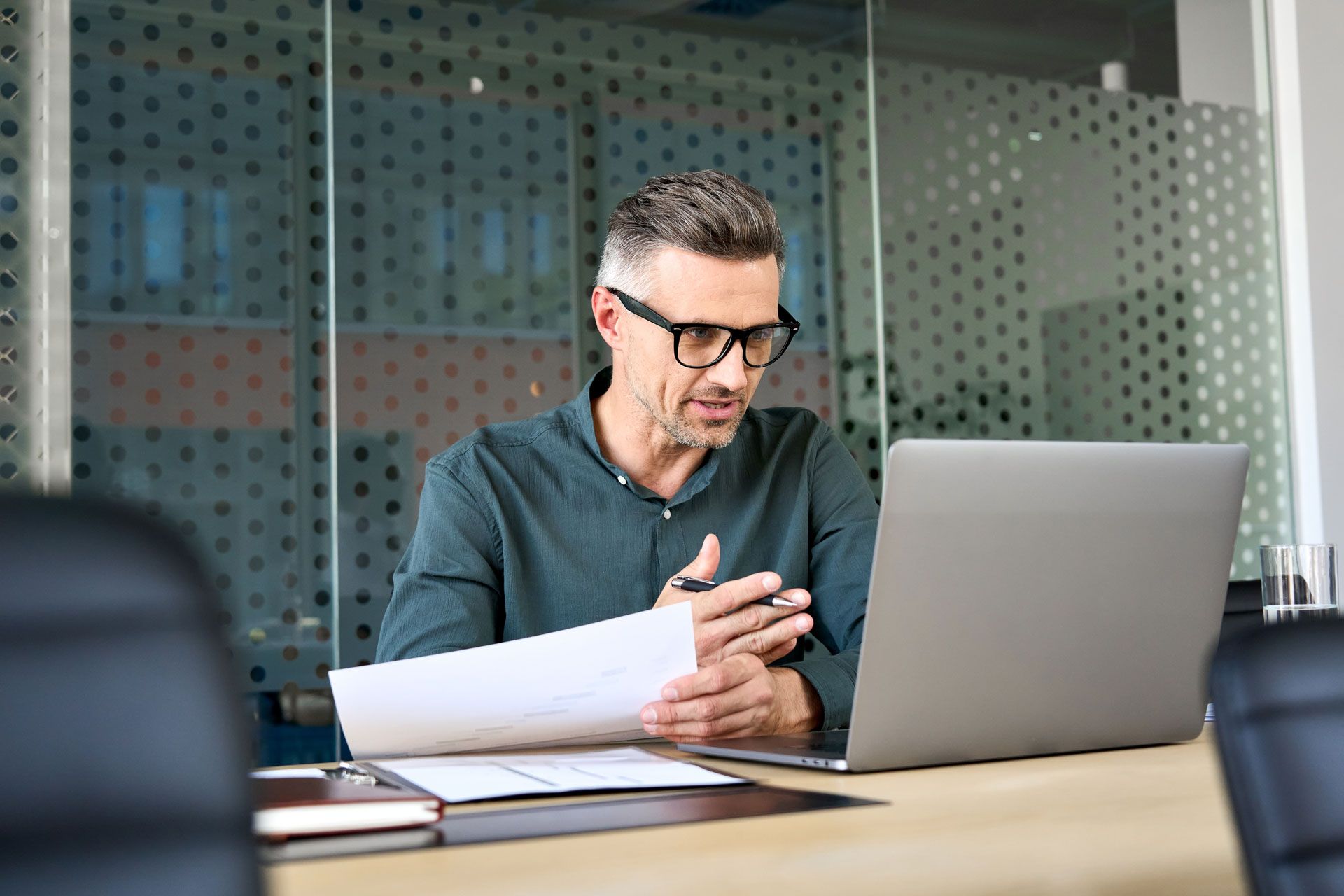 Man with glasses looking at documents, engaged in a video call on a laptop in a modern office.