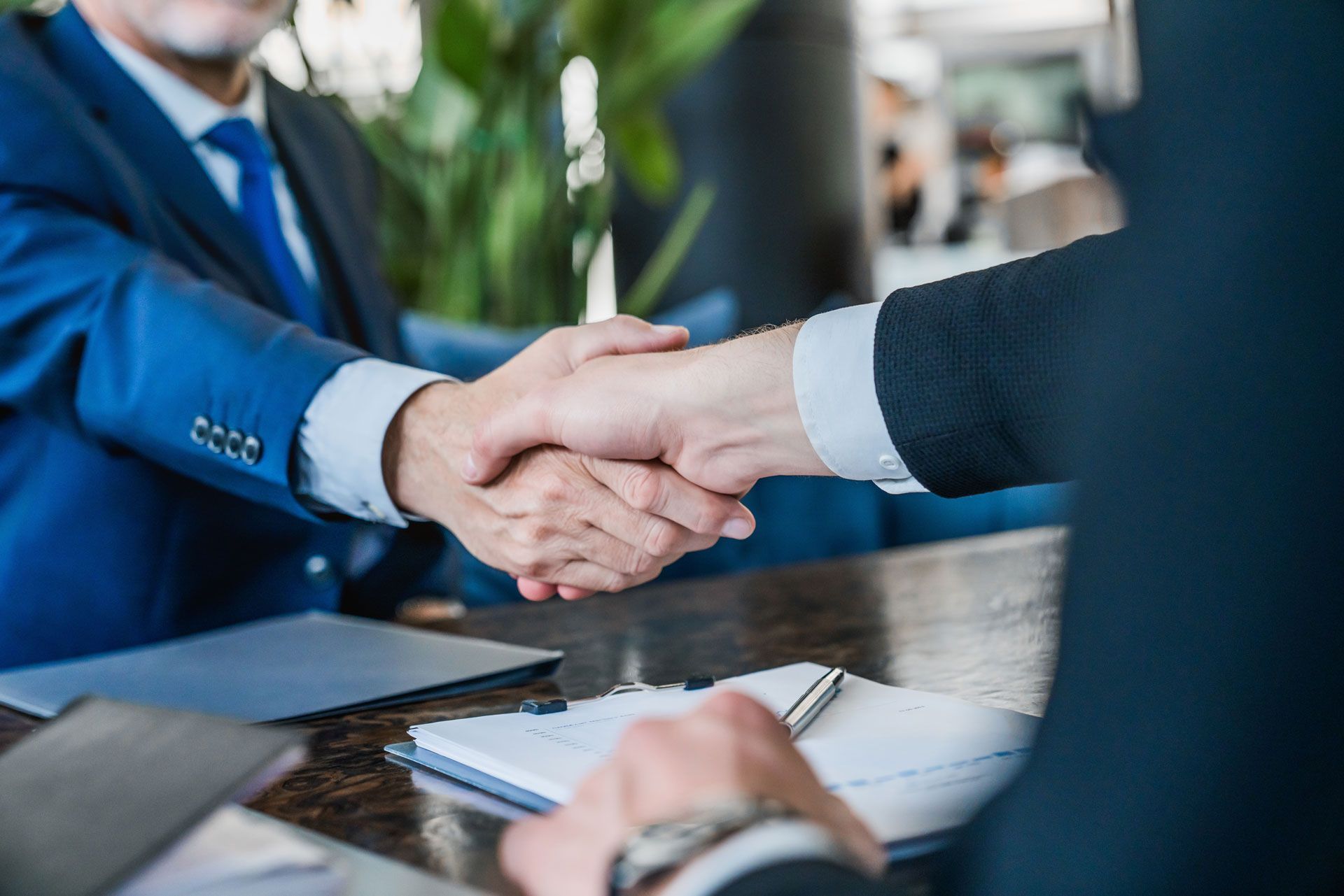 Two men in suits shaking hands over a table with documents, inside a well-lit setting.