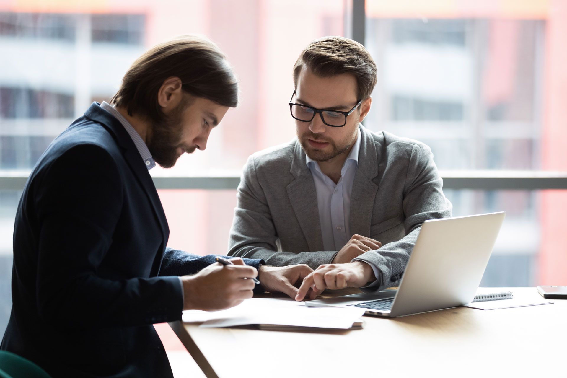 Two men in suits review documents in an office setting; one points at a paper as the other writes.