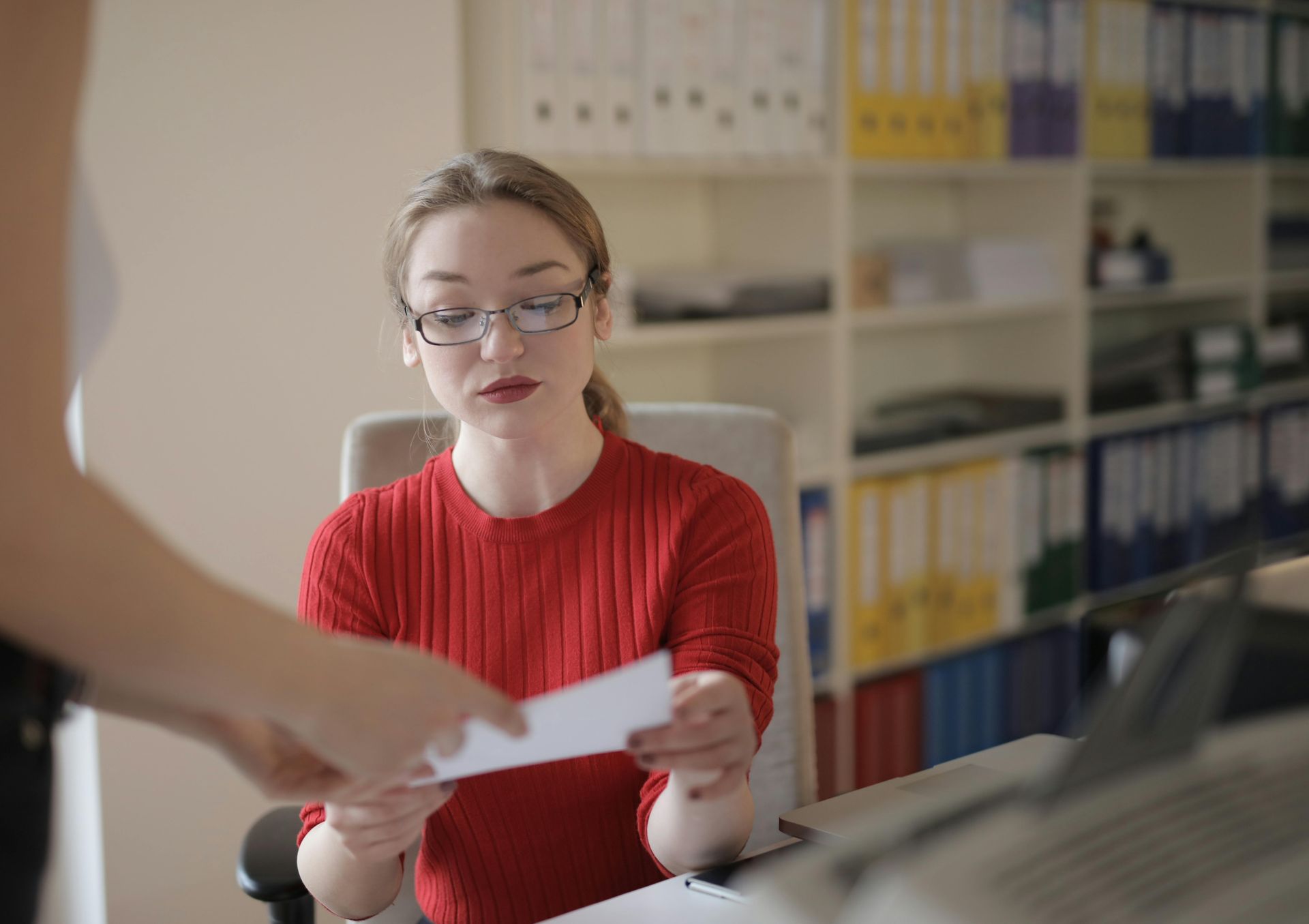 A woman in a red sweater is sitting at a desk holding a piece of paper.