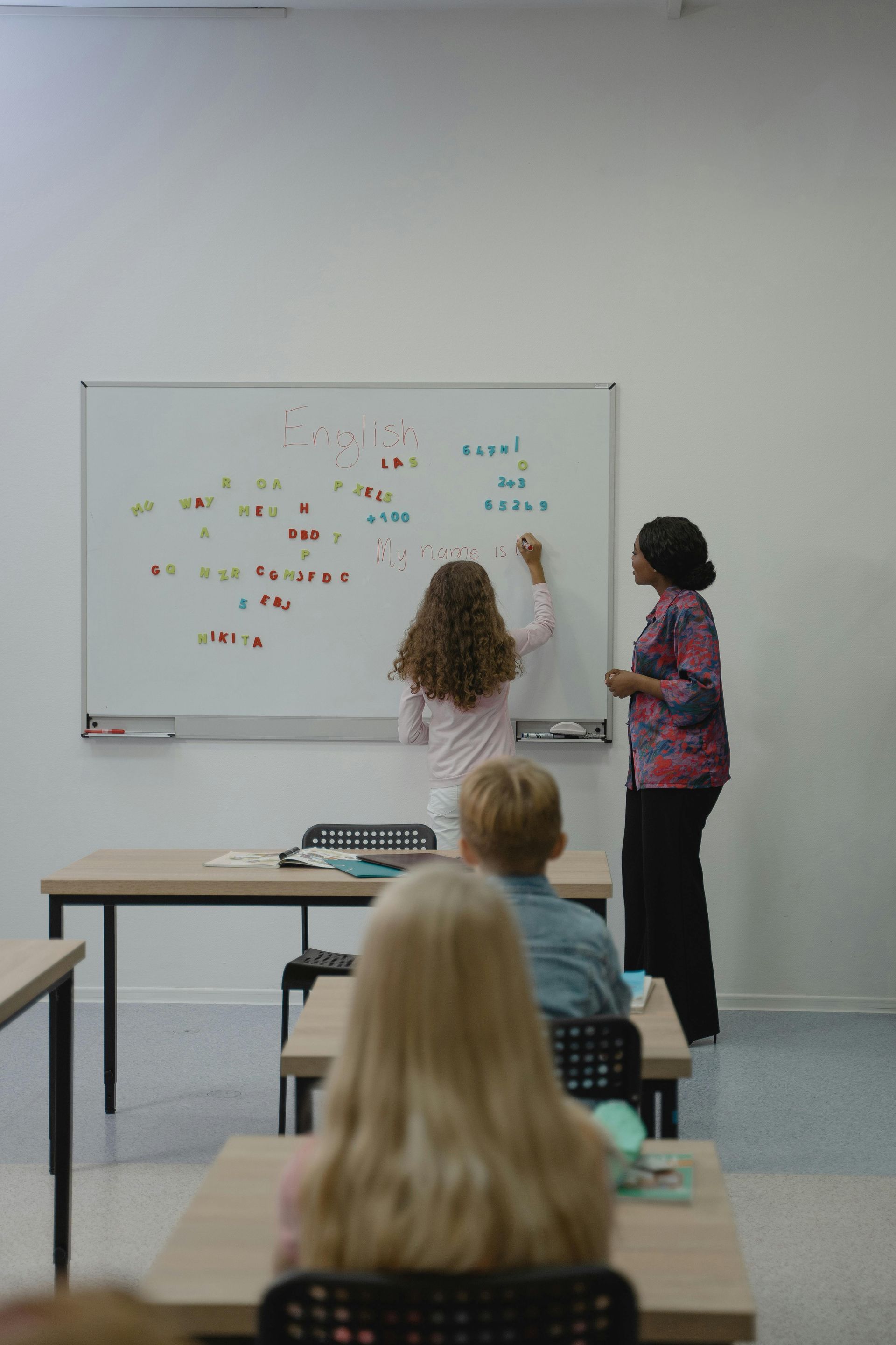 A group of children are sitting at desks in a classroom while a teacher writes on a whiteboard.