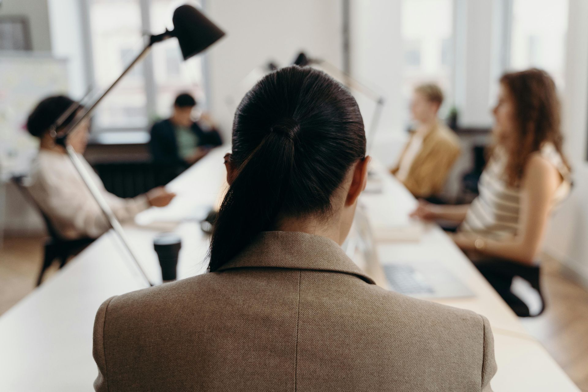 Woman in a tan blazer at the head of a conference table, facing a group of blurred people.