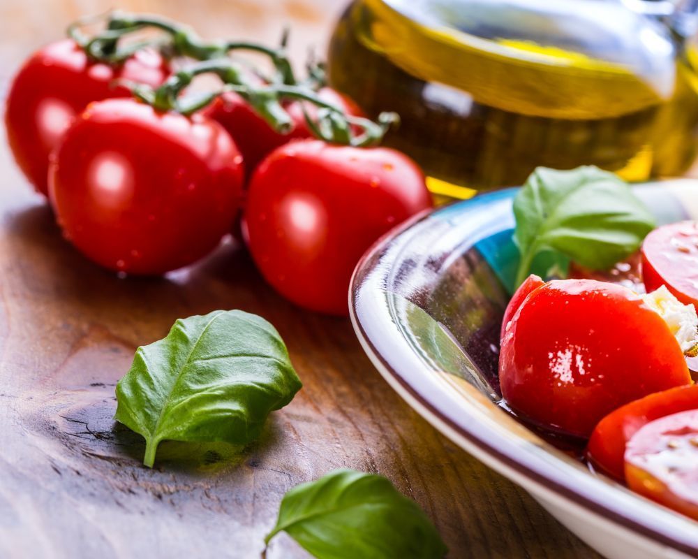 Tomatoes, basil, olive oil, and sliced tomatoes in a bowl, on wooden surface.