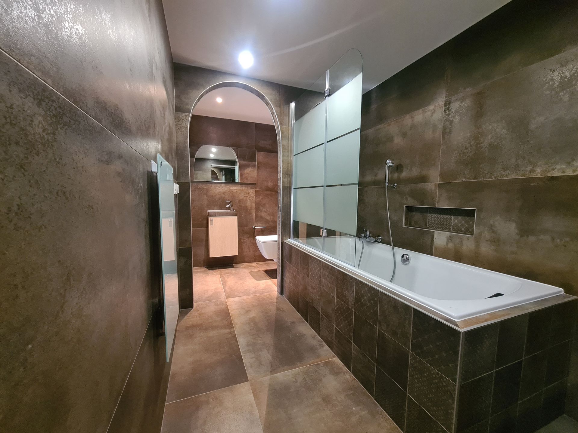 Salle de bains moderne avec murs carrelés de brun, baignoire et porte cintrée.