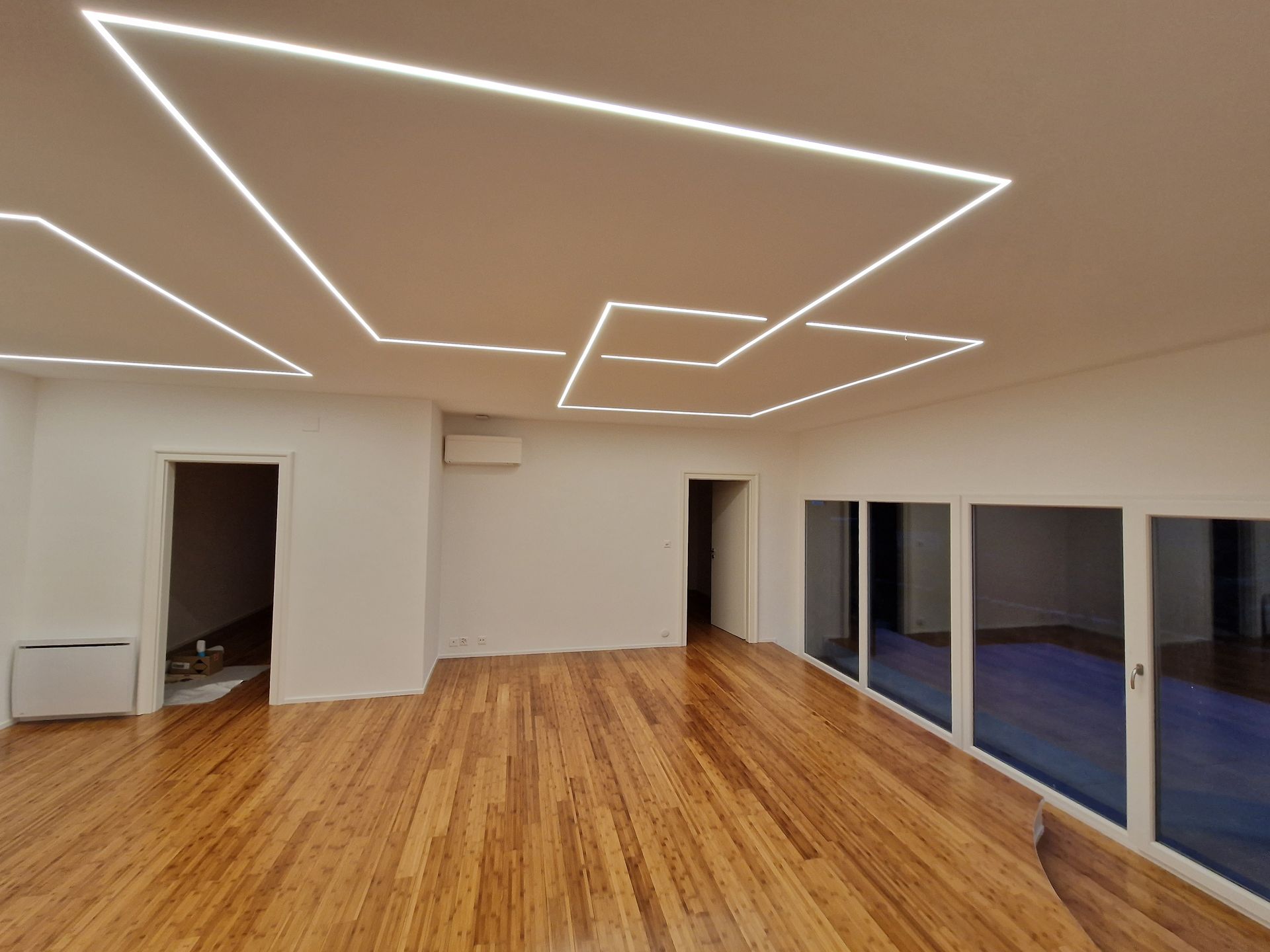 Pièce vide avec parquet, murs blancs et luminaires géométriques au plafond. Portes coulissantes en verre à droite.