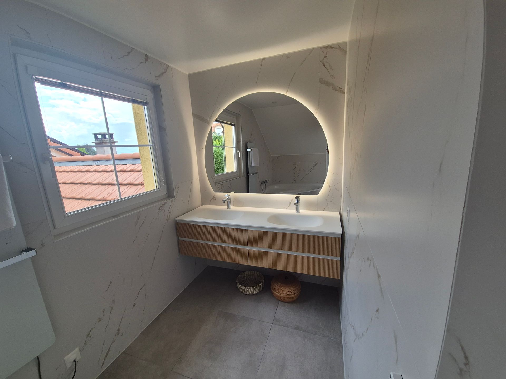 Salle de bains avec miroir rond rétroéclairé, meuble-lavabo et fenêtre. Mur effet marbre, éléments en bois et panier au sol.
