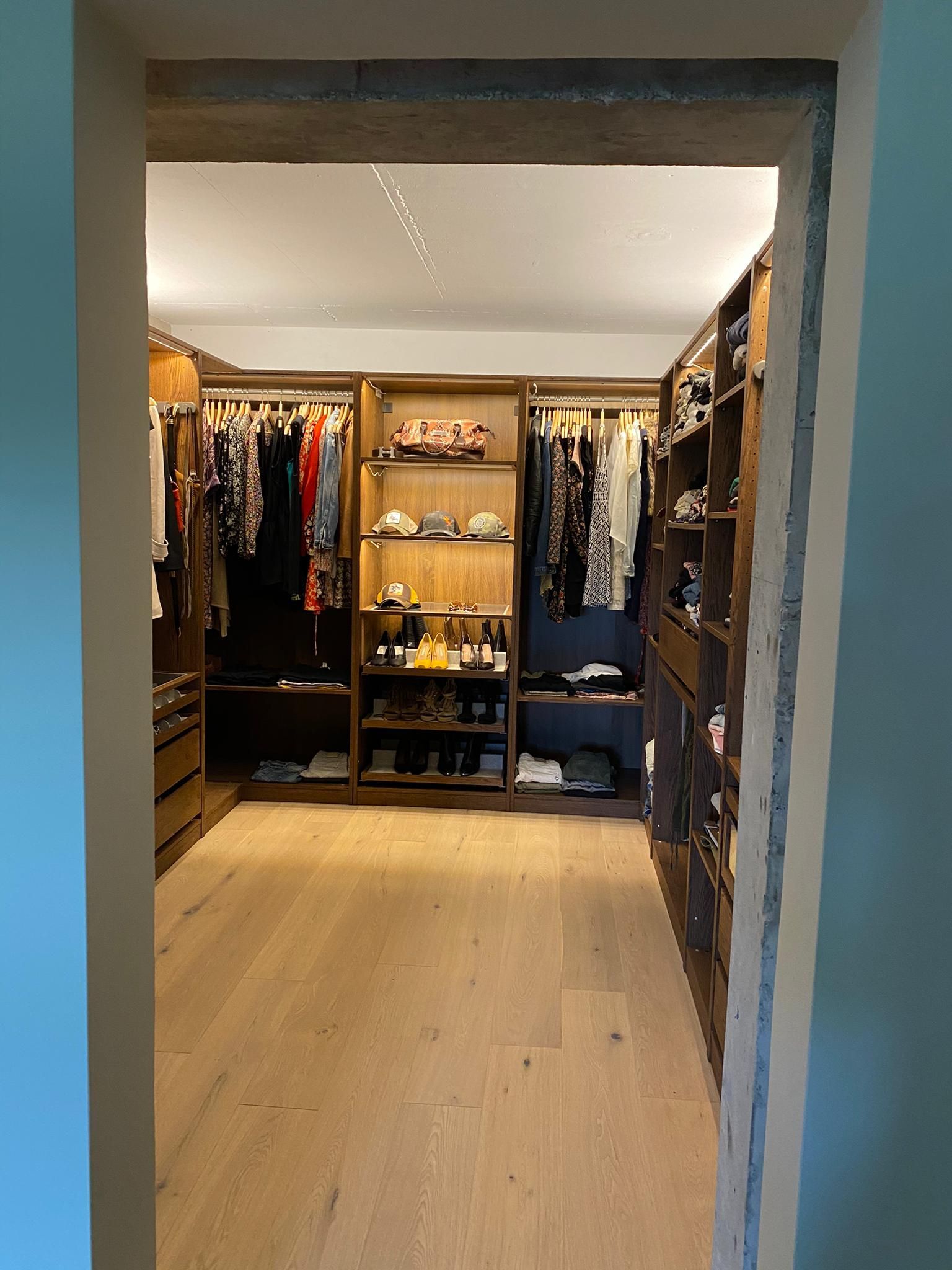 Dressing avec étagères en bois et vêtements. Parquet clair, éclairage encastré. Vue depuis la porte.