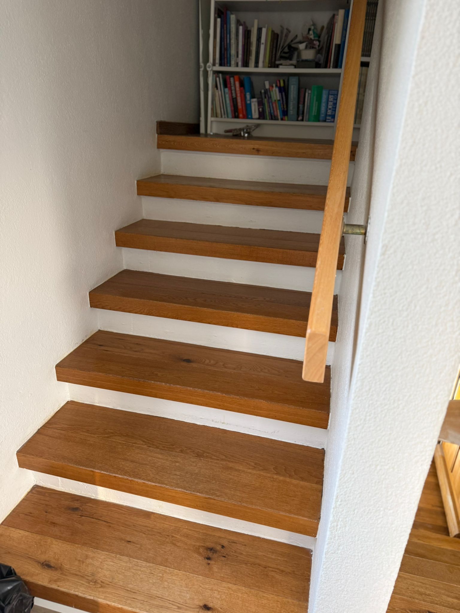Escalier en bois avec contremarches blanches, menant à une bibliothèque. Rambarde à droite.