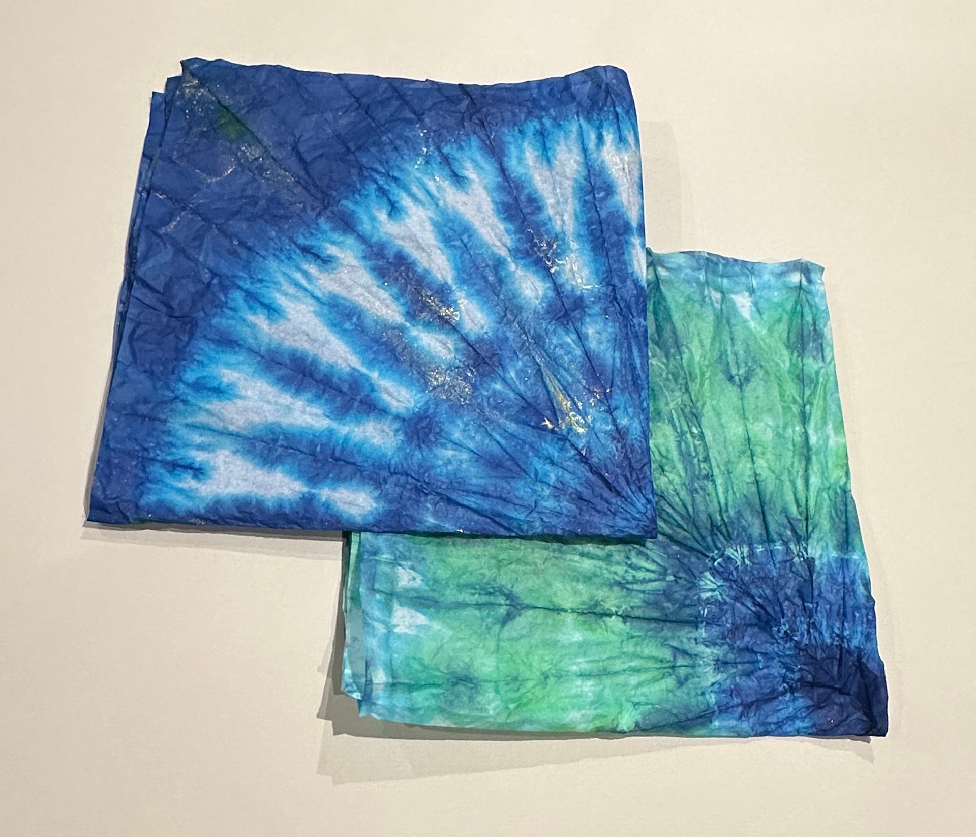 Two blue and green tie dye napkins are sitting on top of each other on a table.