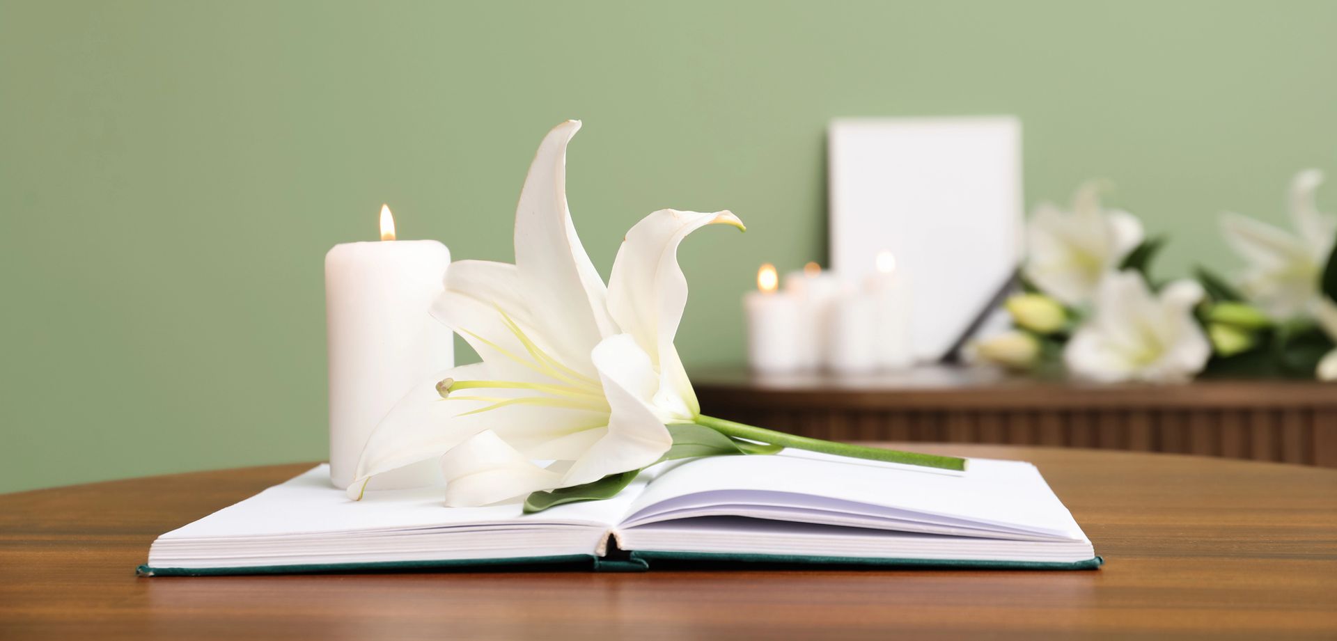 Un giglio bianco e una candela accesa sono appoggiati su un libro aperto su un tavolo di legno, con altre candele e fiori sullo sfondo.