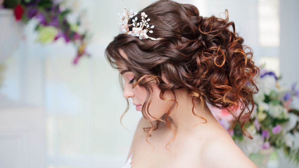 Bride in profile with curled updo and jeweled hairpiece, against a soft floral background