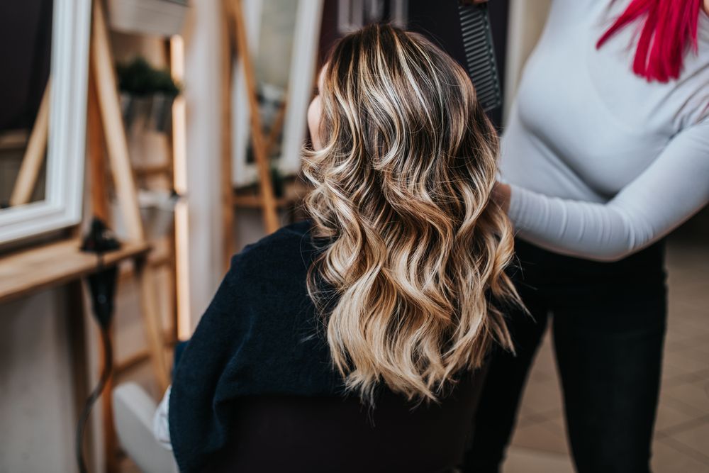 Person with wavy blonde hair seated in a studio while another person adjusts their outfit.