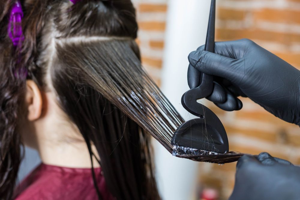 Hair color being applied to a person’s long dark hair with a brush and gloved hands