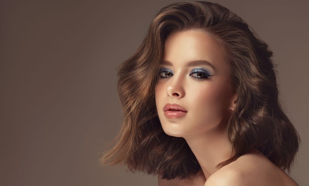 Portrait of a person with short wavy brown hair and soft makeup against a neutral background