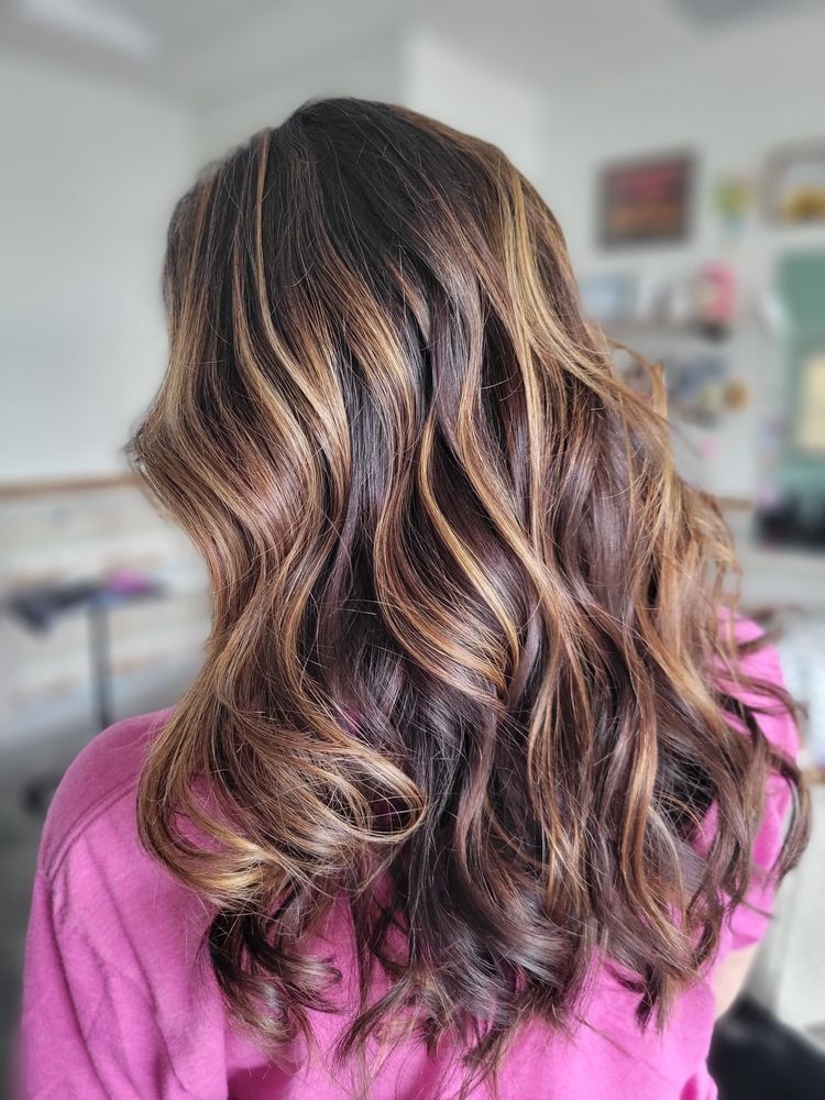 Long wavy brown hair with subtle highlights, shown from behind against a soft indoor background