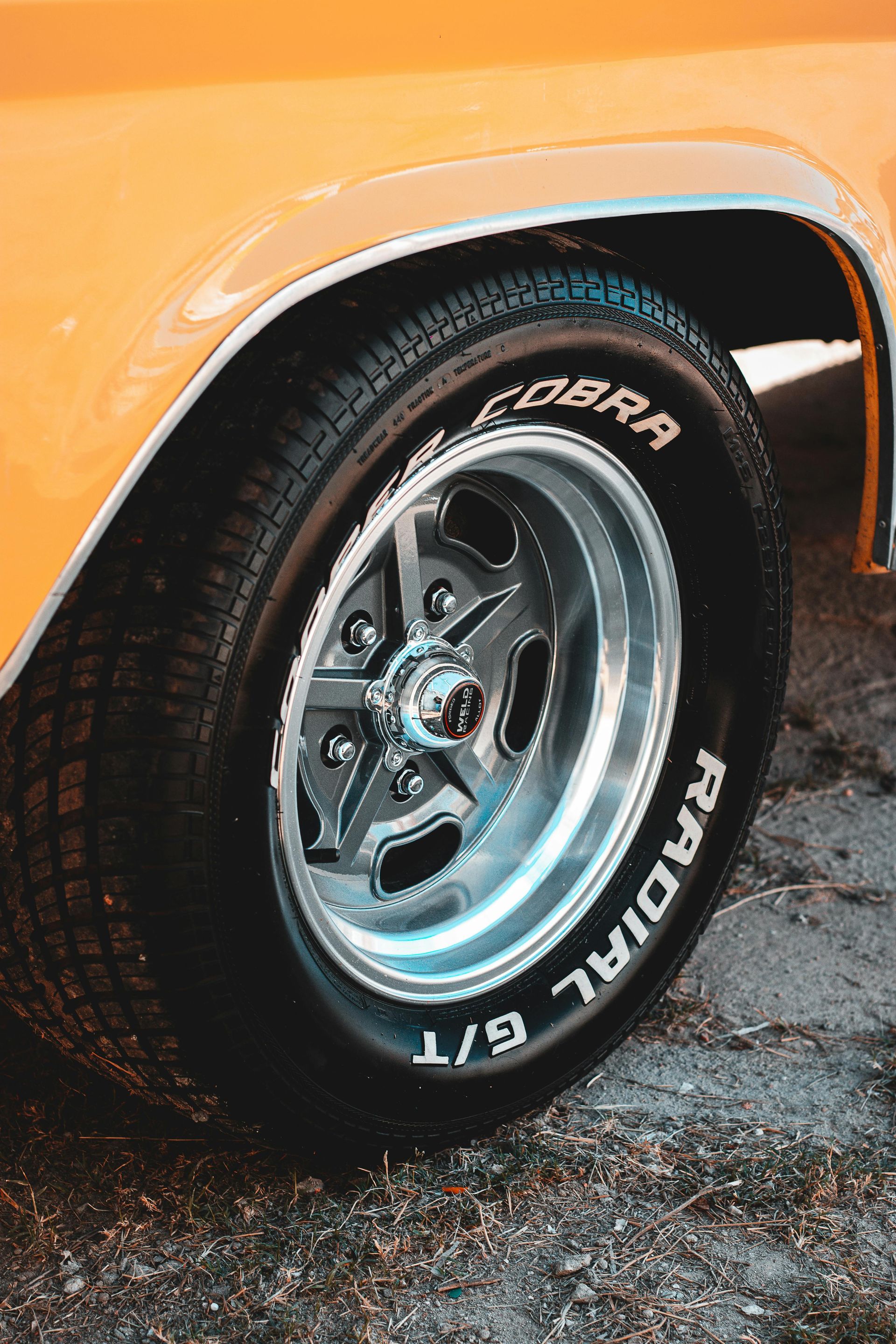 Close-up of an orange car's front tire with a star-shaped chrome rim and white-lettered Cobra Radial G/T tire.