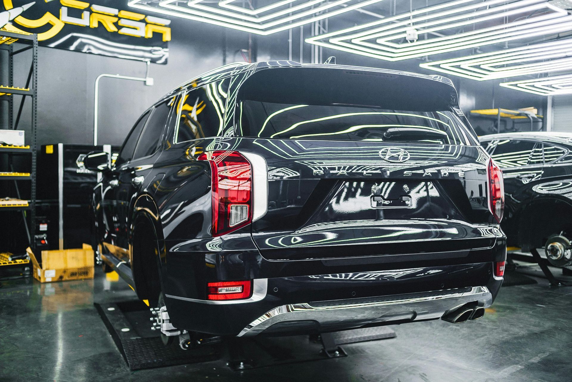 Black SUV in a well-lit garage, seen from the rear. Shiny, with a spoiler, chrome accents, and red taillights.