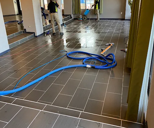 Two people in high-visibility vests clean a tiled floor with professional equipment in an indoor room.