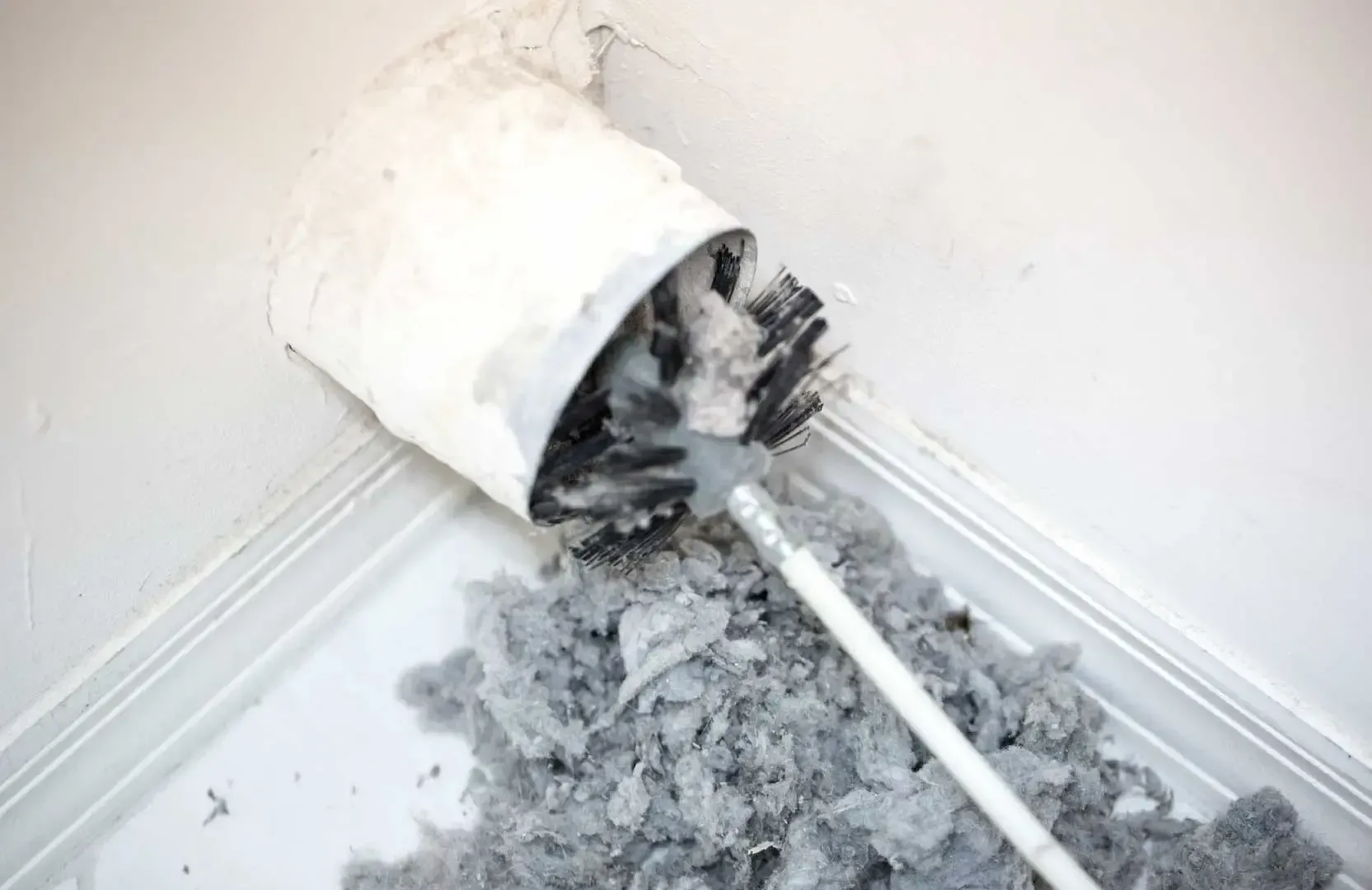 A dryer vent brush removing a large, dense clump of gray lint from a white wall corner.