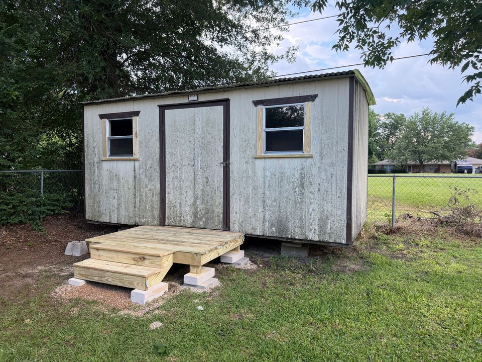 Weathered small shed with two windows and a wooden ramp, sitting in a grassy yard by a fence.
