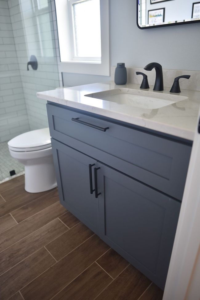 Gray bathroom vanity with sink, black faucet, and toilet beside a window, on wood-look tile floor