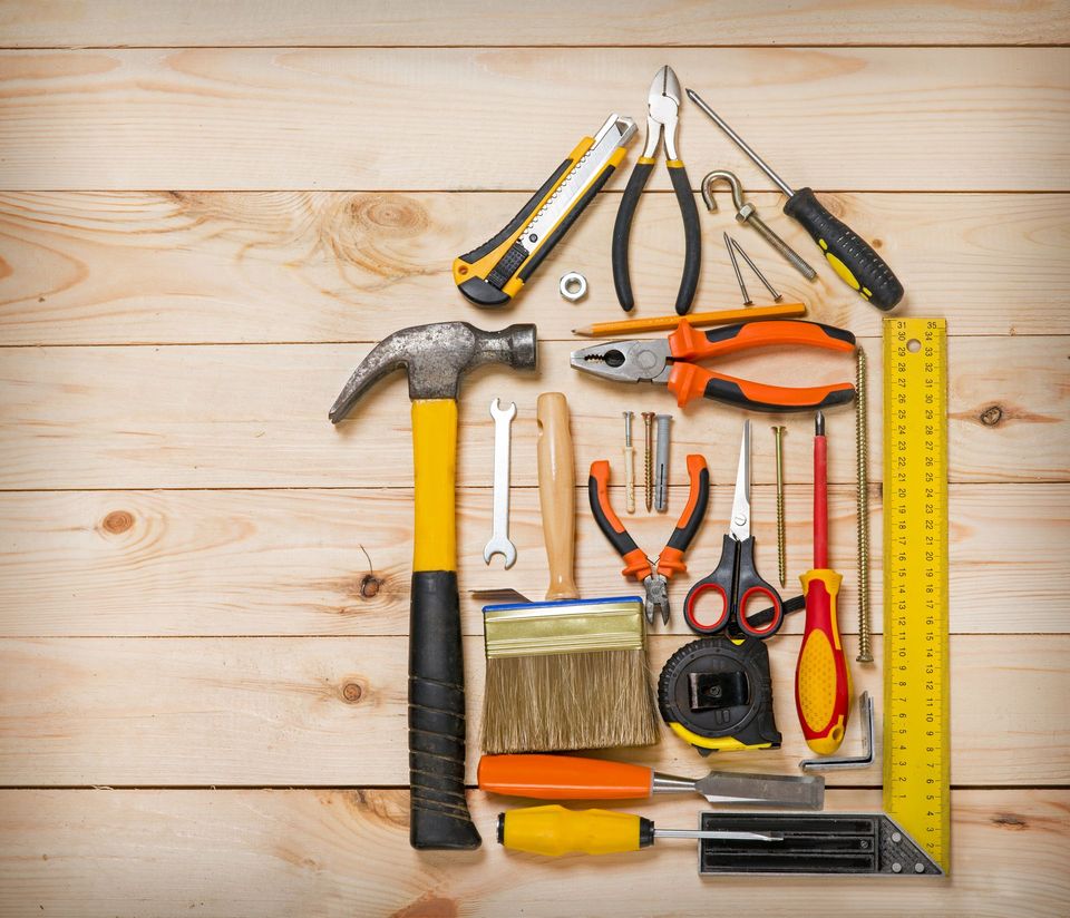 Assorted hand tools arranged on a wooden surface, including hammer, pliers, tape measure, brush, and saw