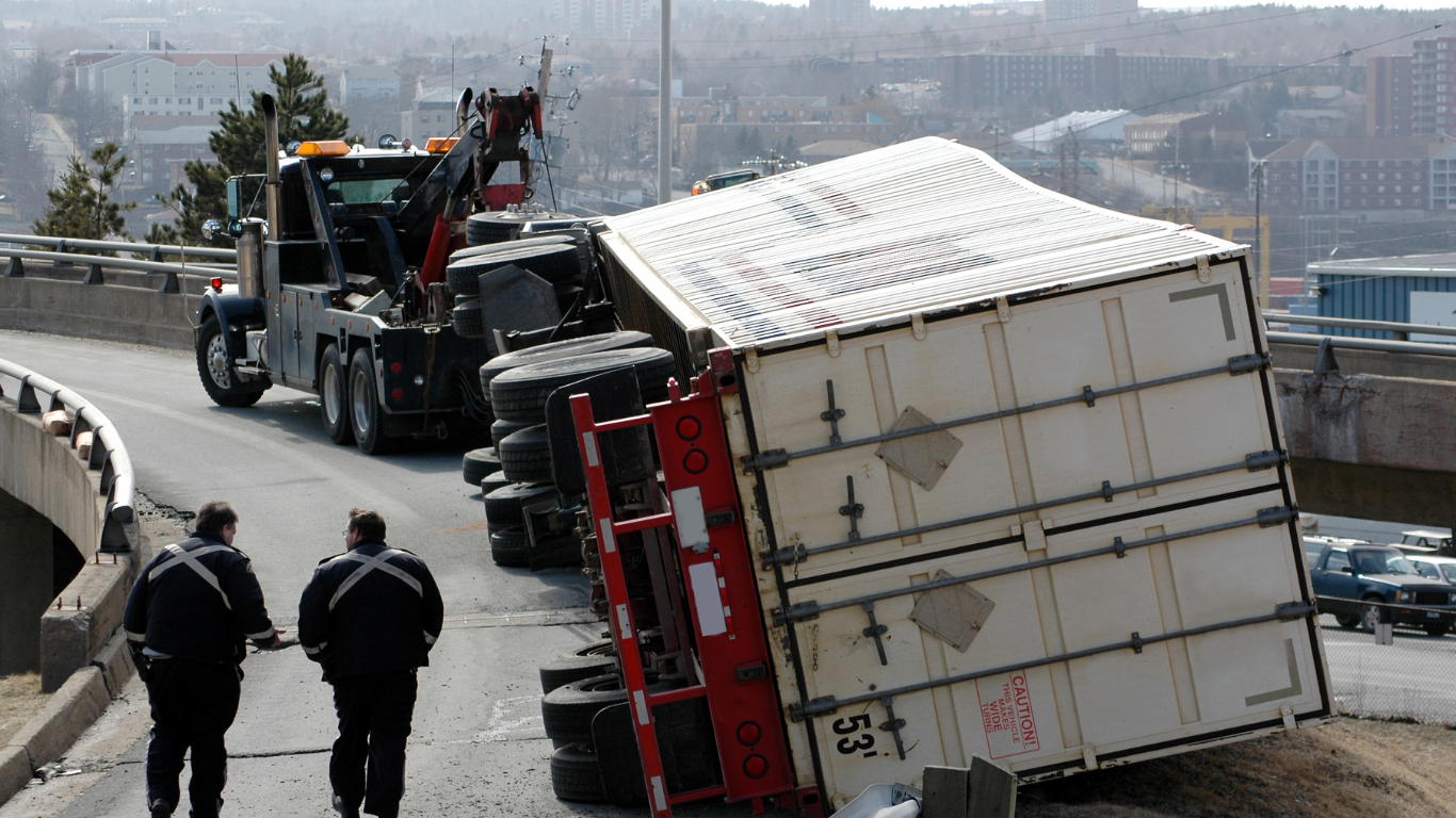 Tow truck attempting to right an overturned semi-truck on a curved overpass. Two people walk in the foreground.