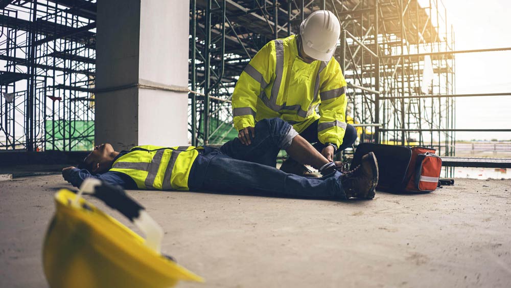 Construction worker assisting an injured colleague on a concrete floor; a first aid kit nearby.