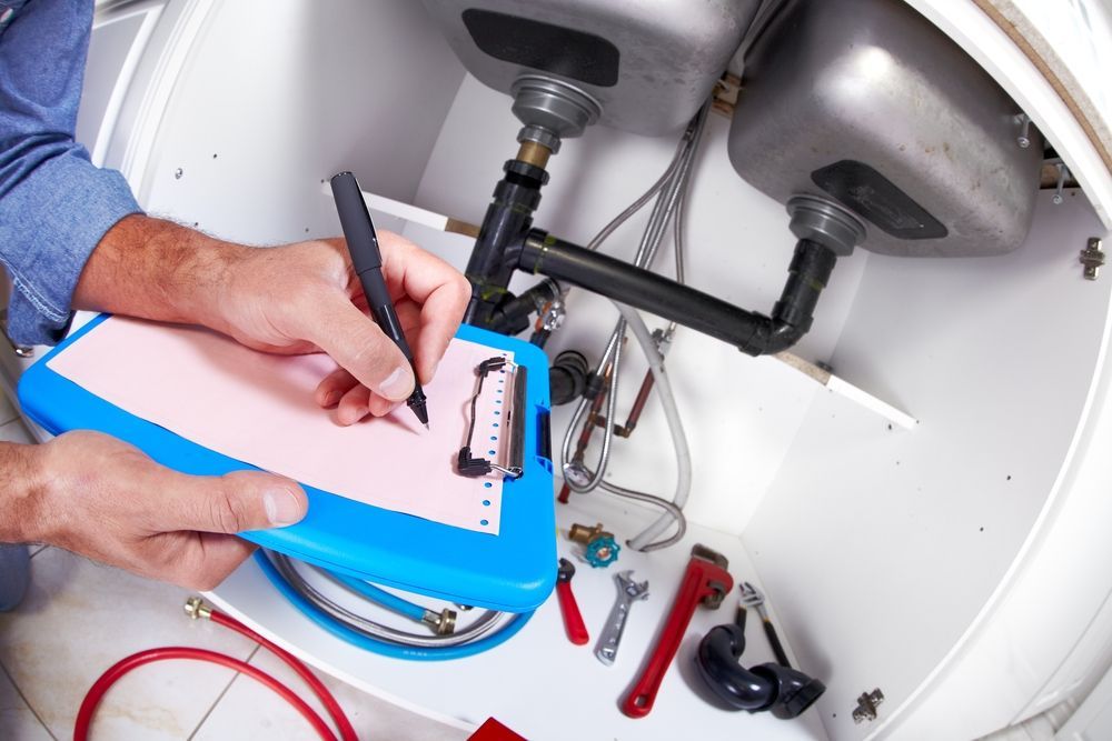 A Plumber Is Writing On A Clipboard Under A Sink — H2O Plumbing & Maintenance Services In Armidale, NSW