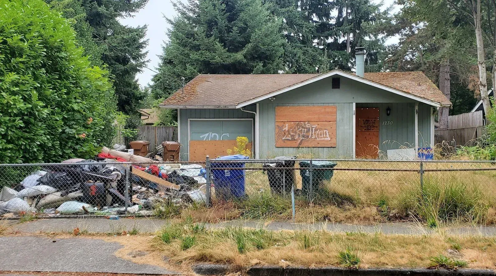 Small house with overgrown yard, trash piles, and black bins by a roadside in a wooded area