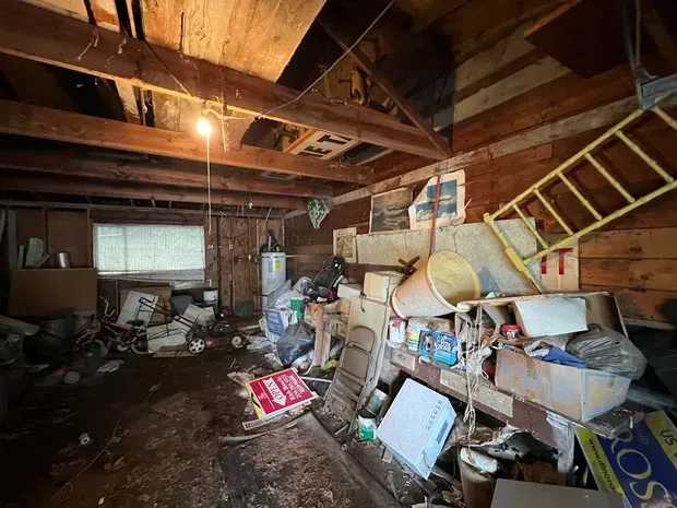 Cluttered attic or garage with junk piles, exposed beams, ladder, and scattered boxes in dim light