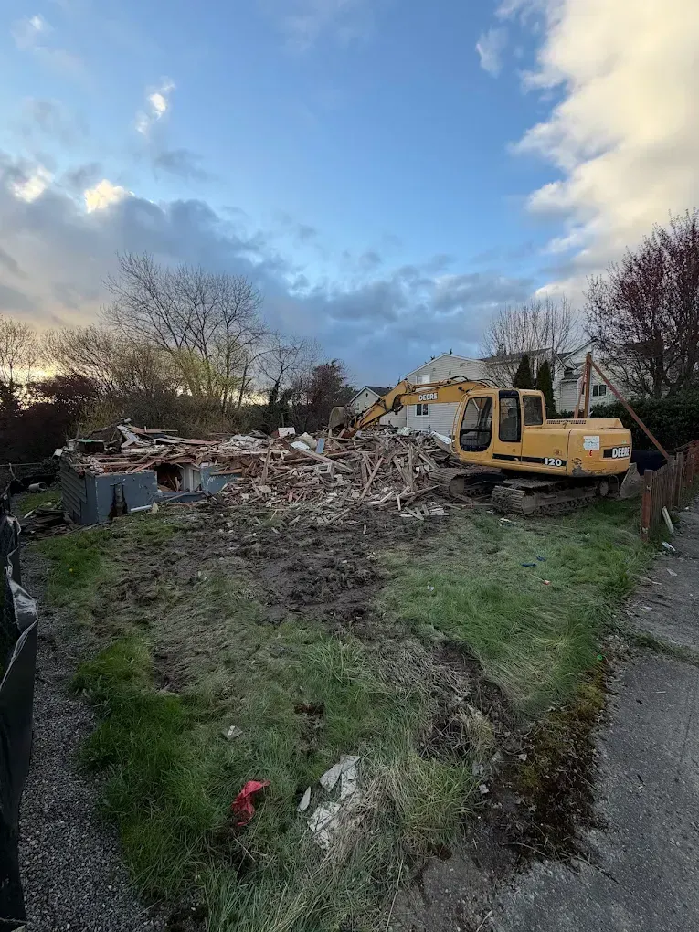 Yellow excavator beside a rubble pile in a muddy yard under a cloudy sky