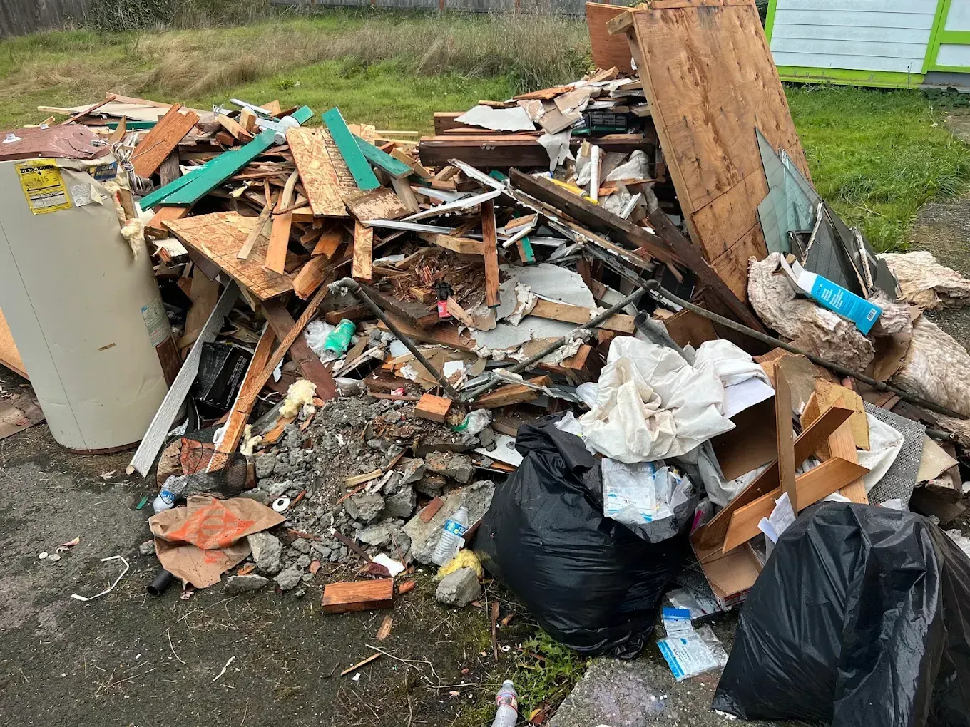 Pile of mixed debris and broken wood with trash bags beside a road on a grassy verge