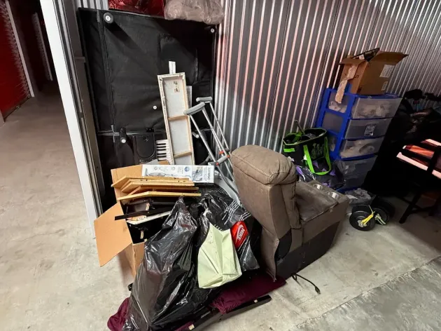 Cluttered storage area with boxed items, tools, and a brown recliner against a corrugated wall