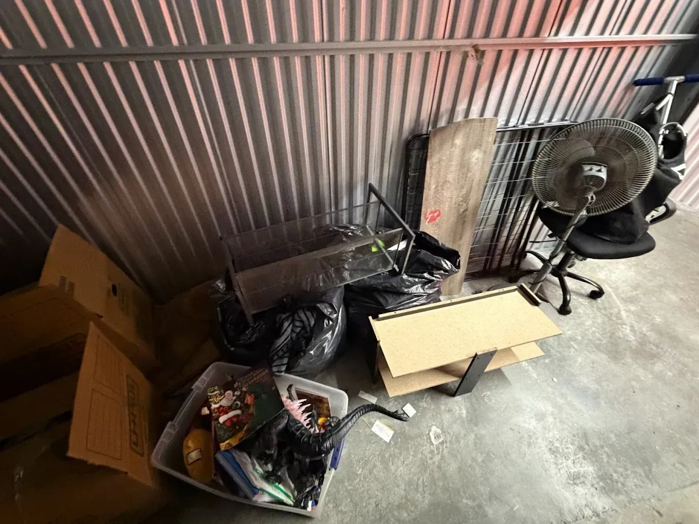 Storage area with boxes, bags, and a fan along a corrugated metal wall on a concrete floor.