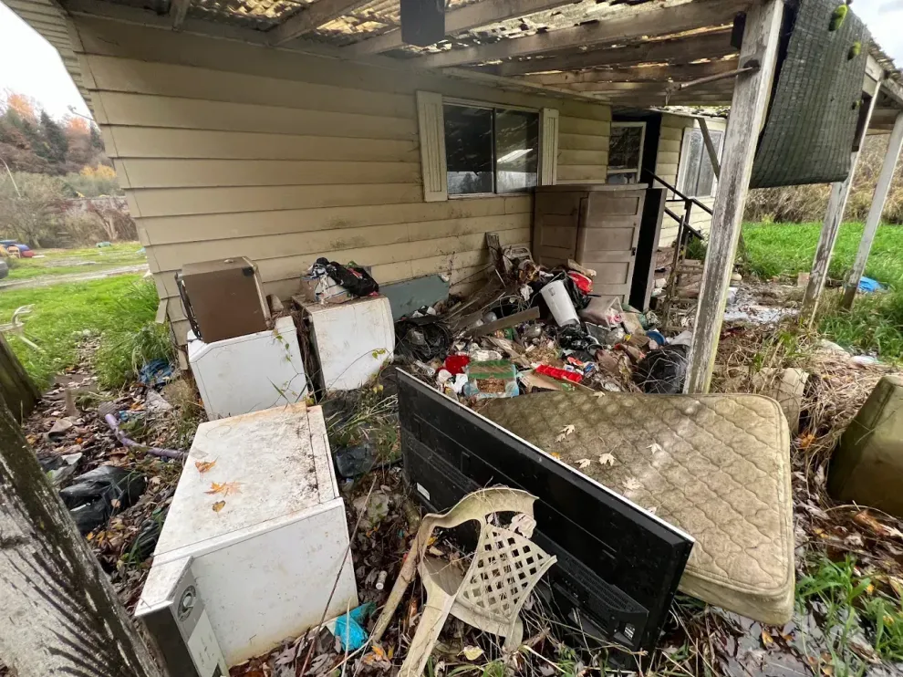 Cluttered porch with debris, boxes, broken furniture, and trash under a damaged roof.