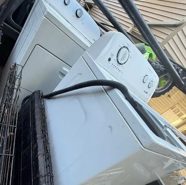 Two stacked white washing machines and a dryer outdoors by a patio roof.