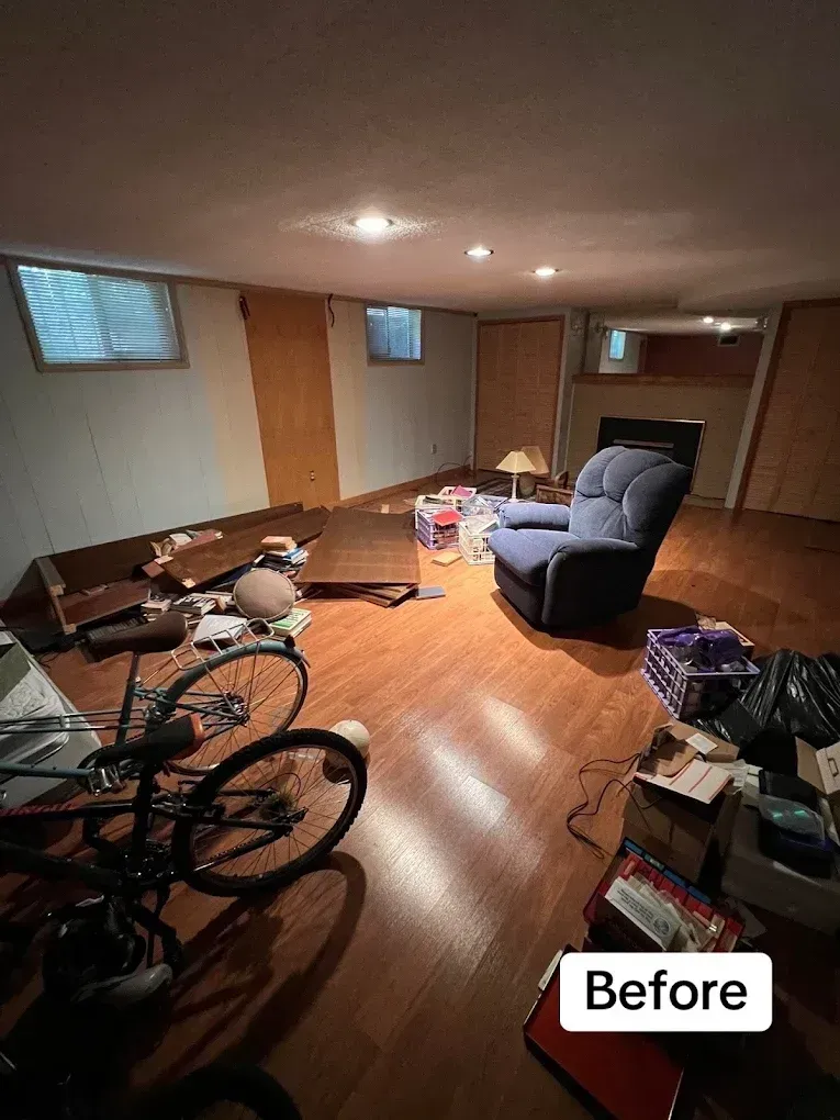 Cluttered basement room with bikes, furniture, boxes, and scattered items on wood flooring