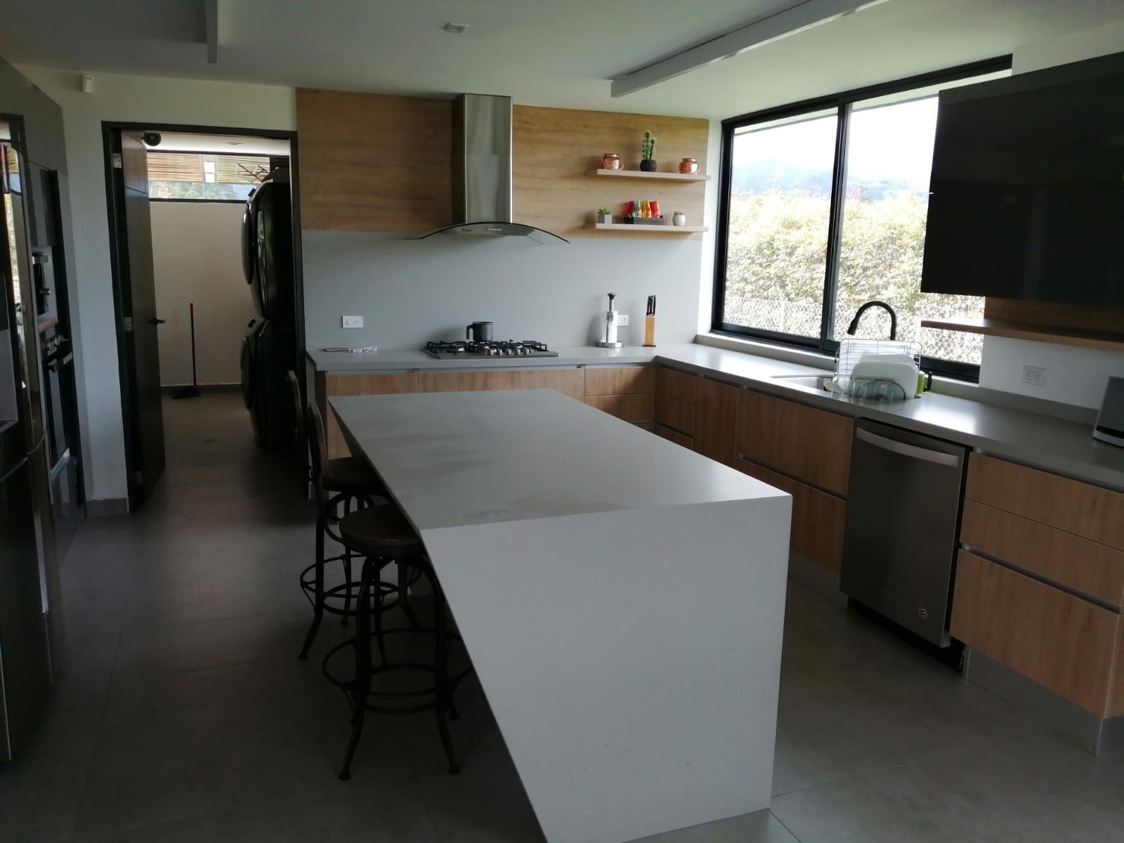 Cocina moderna con isla, armarios de madera, grandes ventanales y fregadero junto a la encimera.