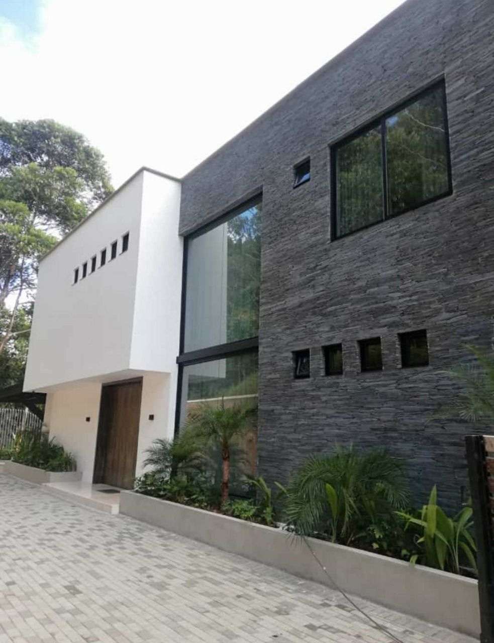 Exterior de casa moderna con fachada de piedra oscura y blanca, grandes ventanales y entrada pavimentada.
