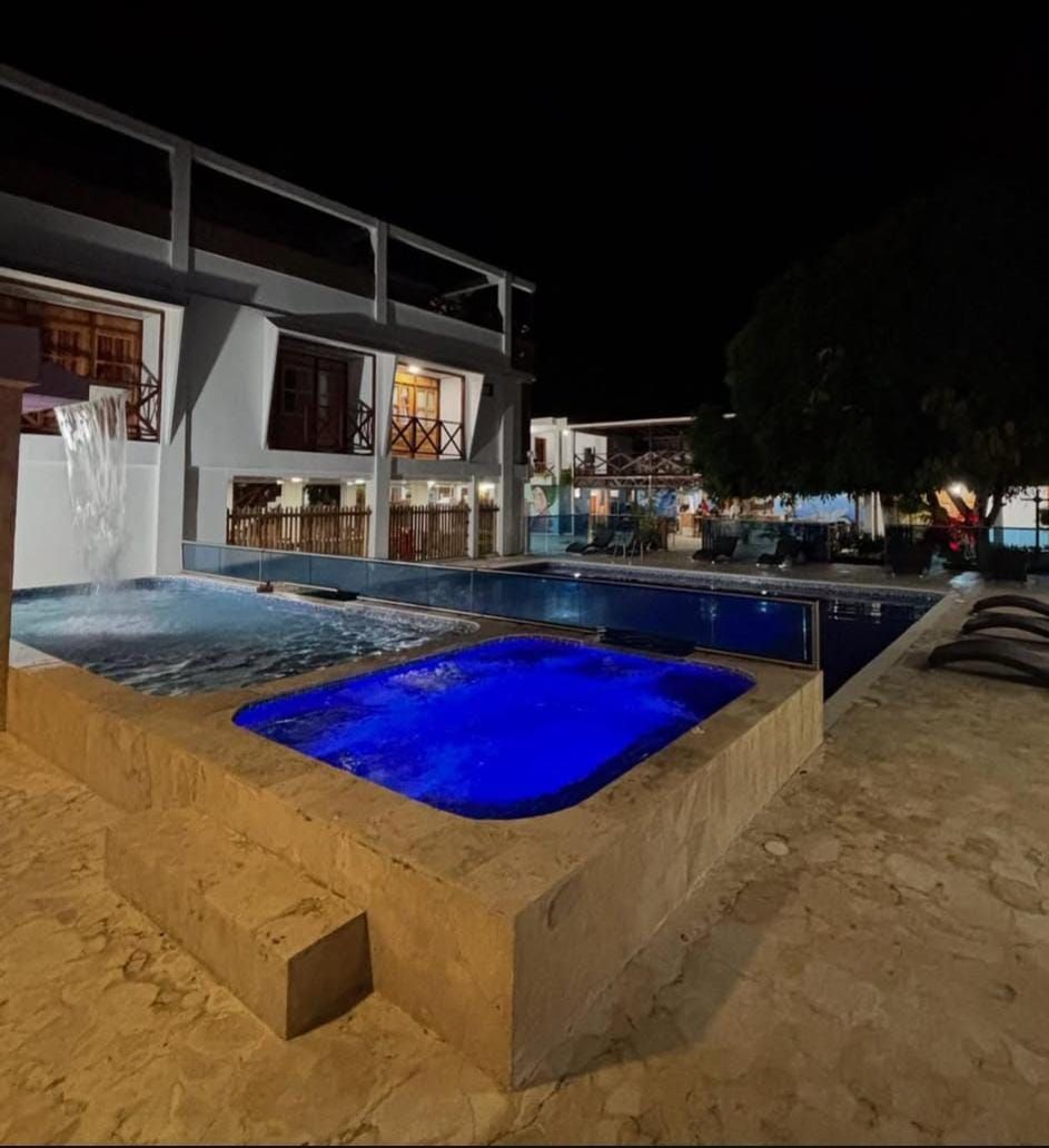 Piscina nocturna tipo resort con agua iluminada con luz azul y tumbonas junto a un edificio de dos plantas.