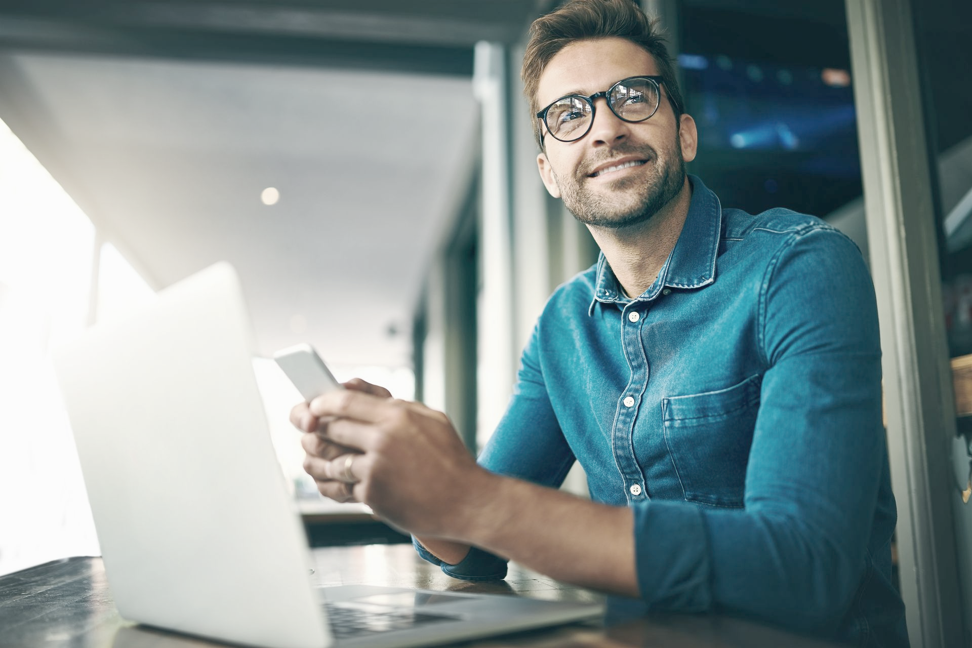 Man with glasses smiling, holding phone, using laptop at work.
