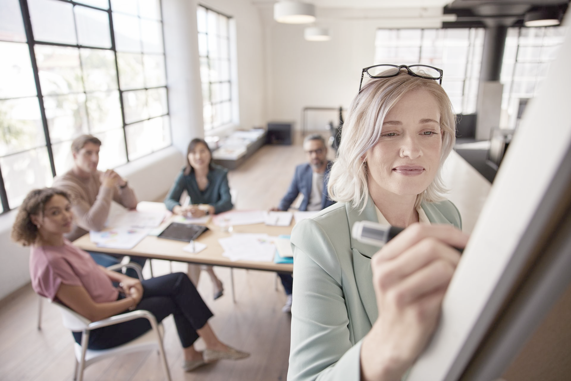 Woman in blazer writes on whiteboard, with colleagues seated at a table in a bright office setting.
