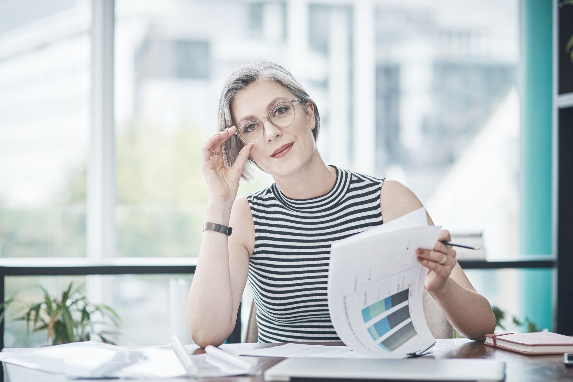 A woman is sitting at a desk holding a piece of paper and adjusting her glasses.