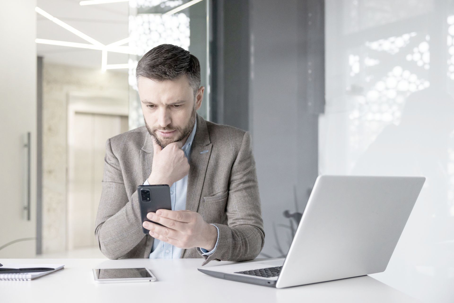 Man in blazer looking intently at his phone, a laptop and second phone on the desk. Modern office setting.